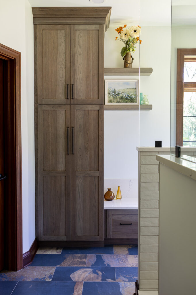 Custom Mouser Cabinetry linen cabinet in Hickory with a Peppercorn stain featuring the Amity door style, built-in storage bench, and floating shelves in a luxury master bathroom remodel in Woodstock, Illinois.